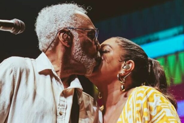 Gilberto Gil, com barba e cabelos grisalhos, beija a testa de sua filha, Preta Gil. Ambos estão em um palco. Gilberto usa uma camisa branca e óculos, enquanto Preta, com os cabelos presos, usa brincos dourados. Um microfone está visível à esquerda.