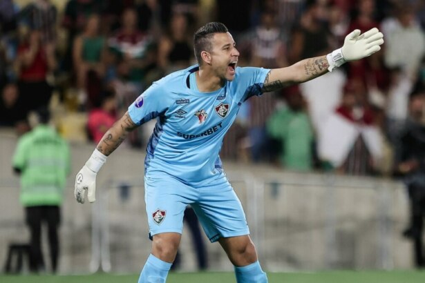 A imagem mostra o goleiro Fábio, do Fluminense, durante uma partida de futebol. Ele usa o uniforme de goleiro na cor azul-claro, com o logo da Umbro e patrocínio da Superbet. Ele tem os braços abertos e as luvas brancas, gritando com uma expressão séria. É possível ver as tatuagens em seus braços. O fundo mostra a arquibancada com torcedores.