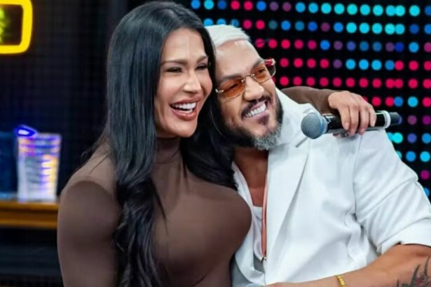 A imagem mostra a atriz e influenciadora Gracyanne Barbosa e o cantor Belo juntos, abraçados, em um estúdio de televisão. Gracyanne sorri, com a cabeça apoiada no ombro de Belo. Ela usa um vestido marrom. Belo também sorri, com a mão direita no ombro de Gracyanne e a esquerda segurando um microfone. Ele usa um casaco branco e óculos de sol com lentes laranjas. Ao fundo, um painel com luzes coloridas e um neon amarelo.