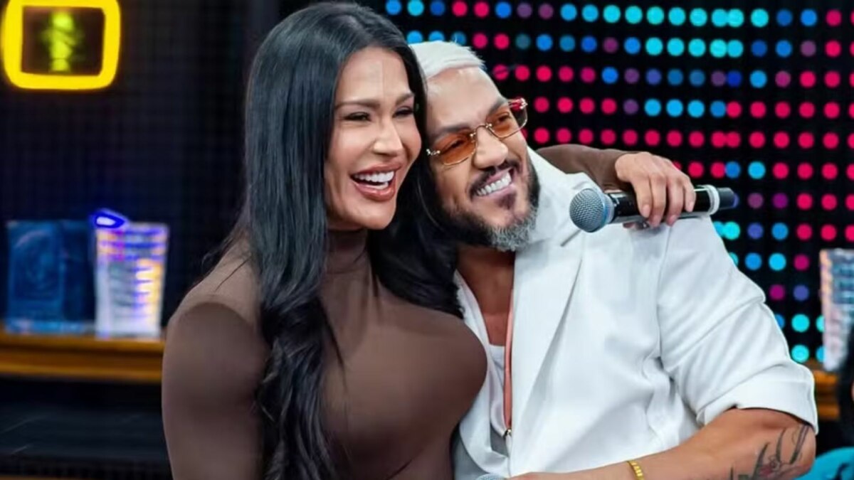 A imagem mostra a atriz e influenciadora Gracyanne Barbosa e o cantor Belo juntos, abraçados, em um estúdio de televisão. Gracyanne sorri, com a cabeça apoiada no ombro de Belo. Ela usa um vestido marrom. Belo também sorri, com a mão direita no ombro de Gracyanne e a esquerda segurando um microfone. Ele usa um casaco branco e óculos de sol com lentes laranjas. Ao fundo, um painel com luzes coloridas e um neon amarelo.