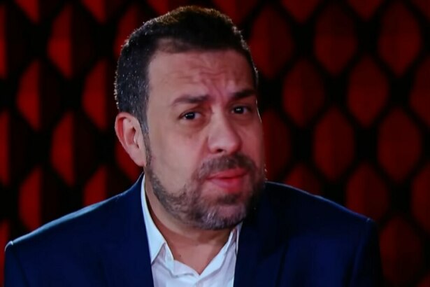 A imagem é um close do político e ativista Guilherme Boulos em uma entrevista para o programa "No Alvo". Ele está olhando para a câmera com uma expressão séria. Boulos usa um terno azul-marinho sobre uma camisa branca. O fundo da imagem é um padrão de losangos em tons de vermelho escuro.