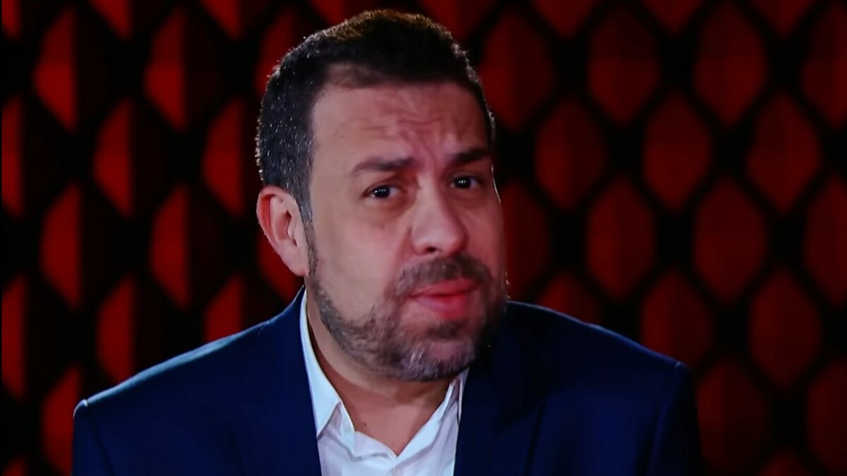 A imagem é um close do político e ativista Guilherme Boulos em uma entrevista para o programa "No Alvo". Ele está olhando para a câmera com uma expressão séria. Boulos usa um terno azul-marinho sobre uma camisa branca. O fundo da imagem é um padrão de losangos em tons de vermelho escuro.