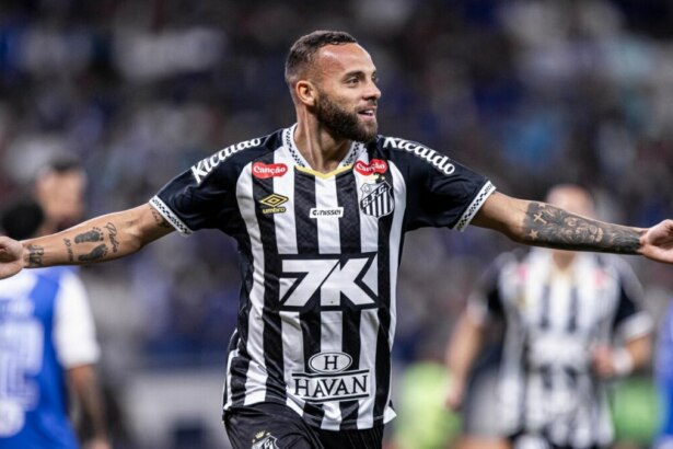 O jogador de futebol Guilherme, do Santos, comemora um gol com os braços abertos. Ele está no gramado, usando a camisa listrada preta e branca do time. Ele tem barba e tatuagens nos braços, e sorri enquanto olha para o lado. O nome do arquivo o associa a um gol em um jogo entre Santos e Cruzeiro.