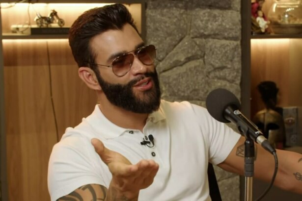 O cantor sertanejo Gusttavo Lima, usando óculos de sol e camisa polo branca, gesticula enquanto fala em um microfone durante um podcast.