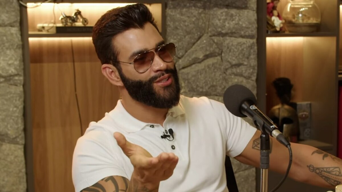 O cantor sertanejo Gusttavo Lima, usando óculos de sol e camisa polo branca, gesticula enquanto fala em um microfone durante um podcast.