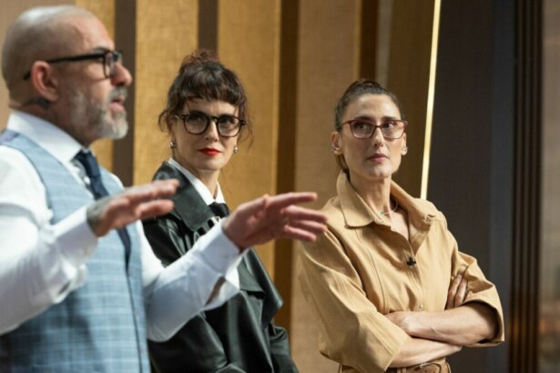 Os três jurados do programa MasterChef Brasil em pé na cozinha do estúdio. Henrique Fogaça, à esquerda, gesticula com as mãos enquanto fala. Helena Rizzo e Paola Carosella, no centro e à direita, respectivamente, o observam com expressões sérias.
