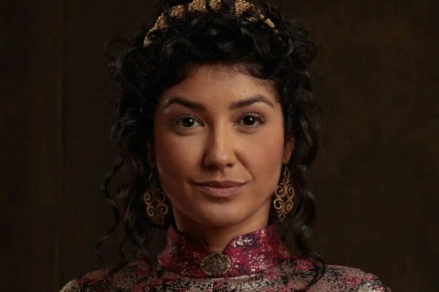A atriz Ingrid Conte em um close-up. Ela tem os cabelos escuros presos, usa uma coroa dourada, brincos grandes e uma blusa com detalhes em vermelho, dourado e preto. Ela olha para a câmera com uma expressão séria.