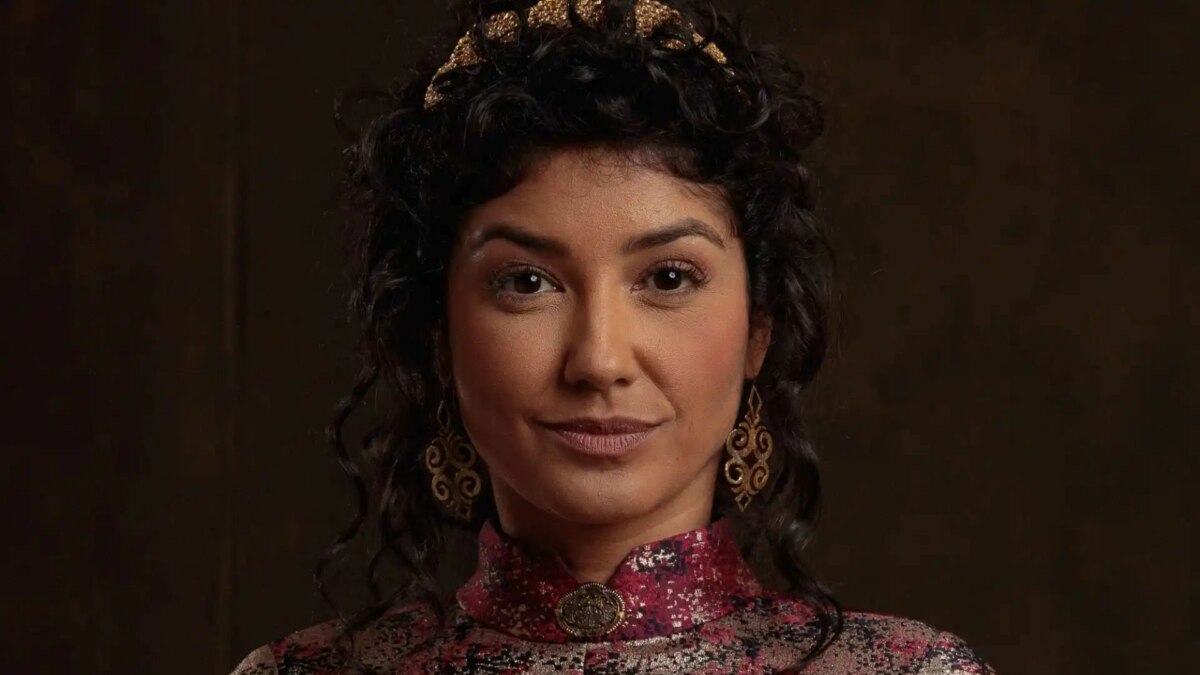 A atriz Ingrid Conte em um close-up. Ela tem os cabelos escuros presos, usa uma coroa dourada, brincos grandes e uma blusa com detalhes em vermelho, dourado e preto. Ela olha para a câmera com uma expressão séria.