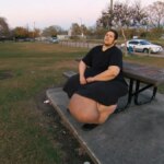 Paciente J.T. do programa "Quilos Mortais", sentado na lateral de um banco de cimento acoplado a uma mesa de piquenique. Ele usa uma camiseta e calças pretas. Sua perna esquerda exibe um grande inchaço causado por linfedema. Ao fundo, há um parque com árvores, grama e uma estrada
