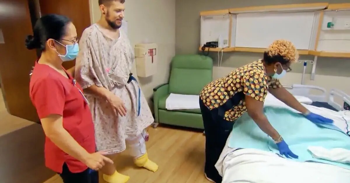 J.T. em um quarto de hospital, sendo acompanhado por duas profissionais de saúde, uma arrumando a cama e a outra observando