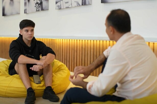 A imagem mostra uma cena do programa "Globo Repórter". À esquerda, um garoto de cabelos escuros está sentado em um pufe amarelo, usando um moletom preto e bermuda preta. Ele tem uma expressão séria e está com as mãos entrelaçadas. À direita, o repórter Jean Raupp está de costas para a câmera, sentado em outro pufe amarelo, com uma camisa social clara. O fundo mostra uma parede com quadros e revestimento de madeira.