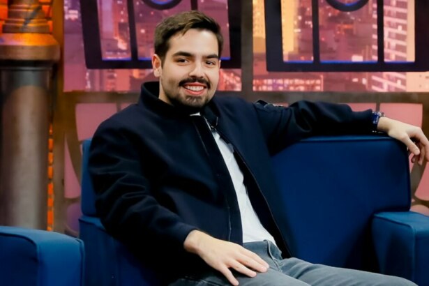 João Silva, convidado do programa 'The Noite com Danilo Gentili', está sentado em uma poltrona azul, sorrindo para a câmera. Ele usa uma jaqueta azul-marinho e o cenário do estúdio, com uma simulação de janelas com a vista de uma cidade à noite, é visível ao fundo.