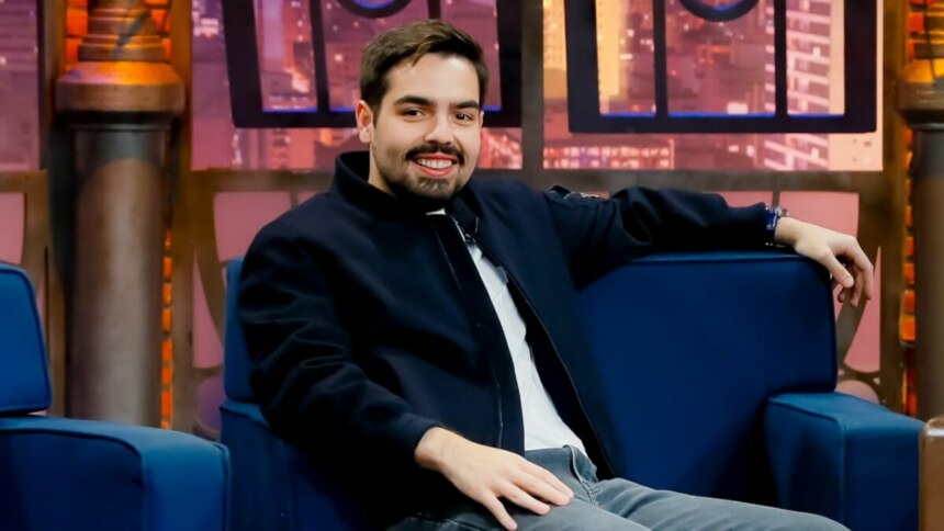 João Silva, convidado do programa 'The Noite com Danilo Gentili', está sentado em uma poltrona azul, sorrindo para a câmera. Ele usa uma jaqueta azul-marinho e o cenário do estúdio, com uma simulação de janelas com a vista de uma cidade à noite, é visível ao fundo.