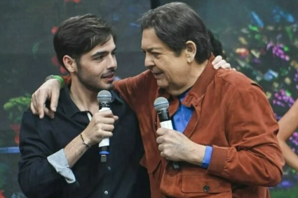 Os apresentadores João Silva e Faustão, pai e filho, em pé no palco de um programa de televisão, seguram microfones e sorriem um para o outro. Faustão, de jaqueta marrom, tem o braço sobre o ombro de João, que veste uma camisa escura. O fundo do palco está desfocado com luzes coloridas.