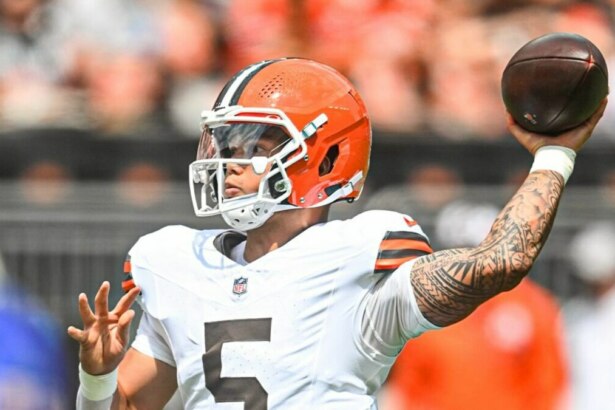 A imagem mostra o jogador de futebol americano Deshaun Watson, do Cleveland Browns, em campo. Ele usa um capacete laranja e branco e tem a bola na mão, prestes a lançá-la. É possível ver as tatuagens em seu braço direito. O fundo da imagem está desfocado, com a arquibancada do estádio.