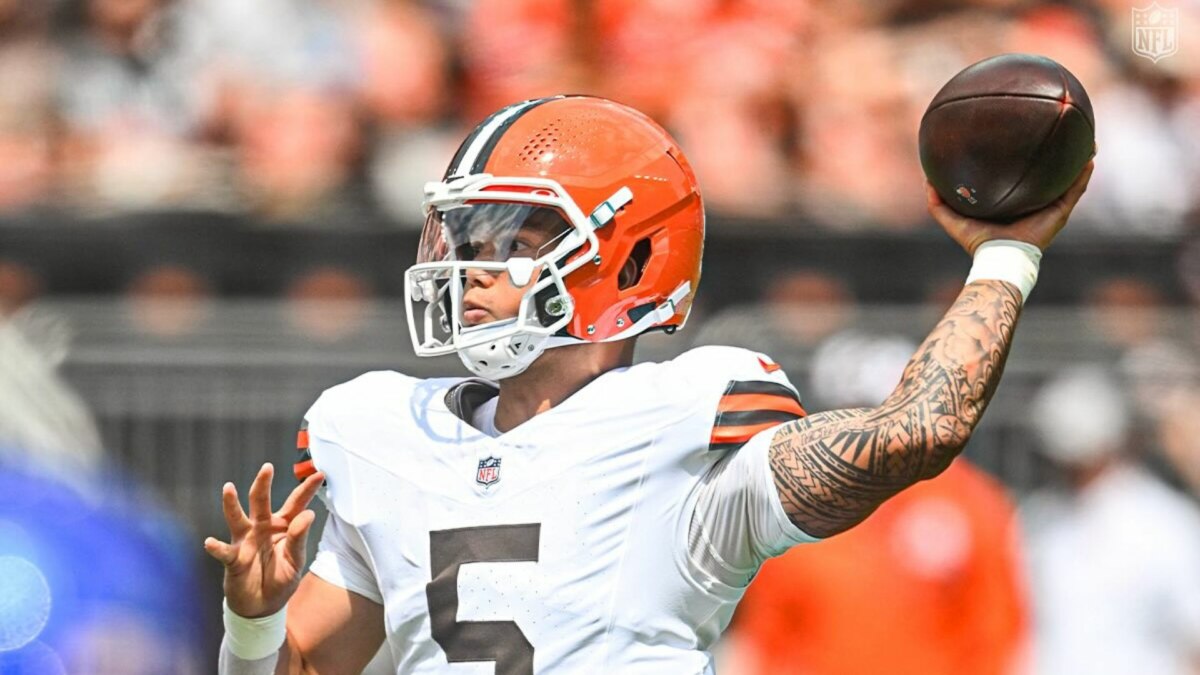 A imagem mostra o jogador de futebol americano Deshaun Watson, do Cleveland Browns, em campo. Ele usa um capacete laranja e branco e tem a bola na mão, prestes a lançá-la. É possível ver as tatuagens em seu braço direito. O fundo da imagem está desfocado, com a arquibancada do estádio.