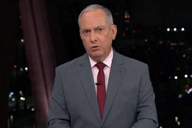 O jornalista José Roberto Burnier, usando um terno cinza, camisa rosa clara e gravata vermelha, apresenta o jornal. Ele olha seriamente para a câmera, com um fundo desfocado que mostra o cenário do telejornal "SP2" e as luzes de uma cidade à noite.