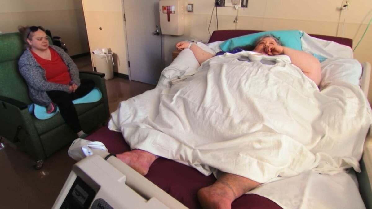 A participante Joyce, do programa Quilos Mortais, deitada em uma cama de hospital, enquanto outra mulher senta ao lado, observando-a com preocupação.