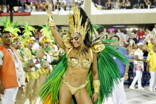 A atriz Juliana Paes, vestida com uma fantasia exuberante de rainha de bateria, nas cores dourado e verde, dança com os braços erguidos durante um desfile de carnaval. Ela sorri com os olhos fechados. Ao fundo, é possível ver outras pessoas fantasiadas e a plateia do sambódromo.