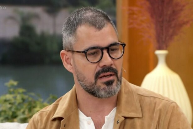 O ator Juliano Cazarré em um close-up, sentado no sofá do programa 'Encontro'. Ele usa óculos de armação escura, uma jaqueta marrom clara e uma camisa branca. Ele tem barba e bigode grisalhos e parece estar falando seriamente. Ao fundo, o cenário do estúdio é visível.