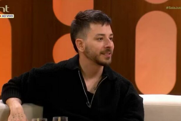 O cantor e apresentador Junior Lima sentado em um sofá no estúdio do programa "Saia Justa". Ele usa um suéter preto de zíper, sorri discretamente e olha para o lado. O logo da emissora GNT está visível no canto superior da tela.