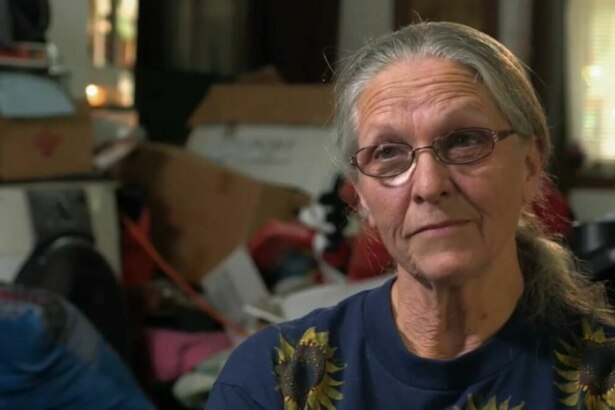 Mulher de cabelos grisalhos e óculos, com uma camiseta de girassol, olha para a câmera com um sorriso sutil. O fundo está desfocado e mostra um ambiente bagunçado, com caixas e objetos empilhados, em trecho do programa Acumuladores