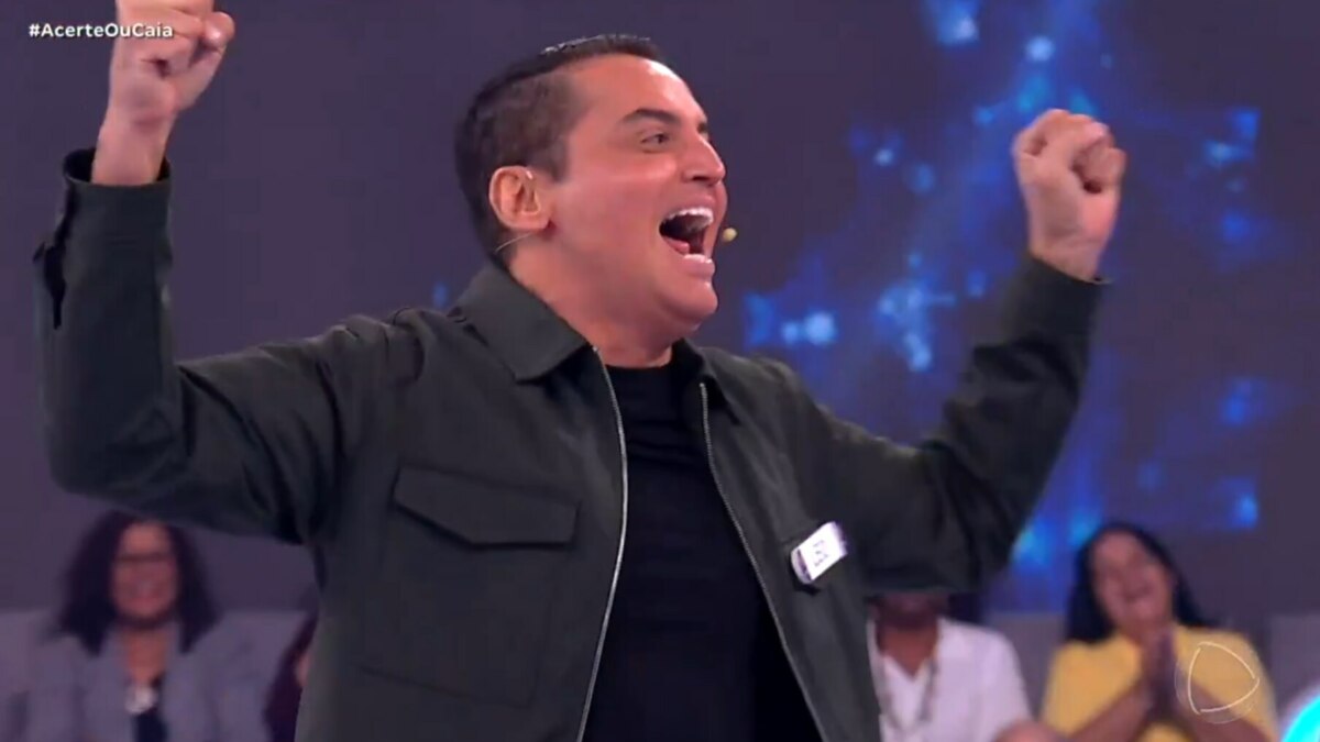 O apresentador Leo Dias, com uma jaqueta verde-escura, celebra com os punhos para o ar em um estúdio do Acerte ou Caia. Ele está sorrindo.