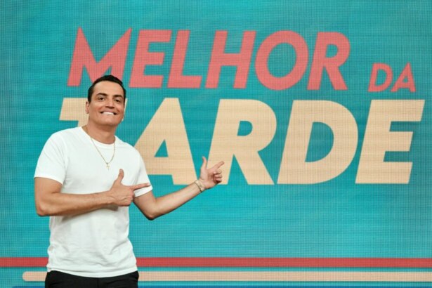 A imagem mostra o apresentador Léo Dias, sorrindo e usando uma camiseta branca e calças pretas. Ele aponta com as duas mãos para um painel com a inscrição "MELHOR DA TARDE" em letras maiúsculas vermelhas, sobre um fundo azul-petróleo. A foto foi tirada em um estúdio de televisão e a iluminação é clara.