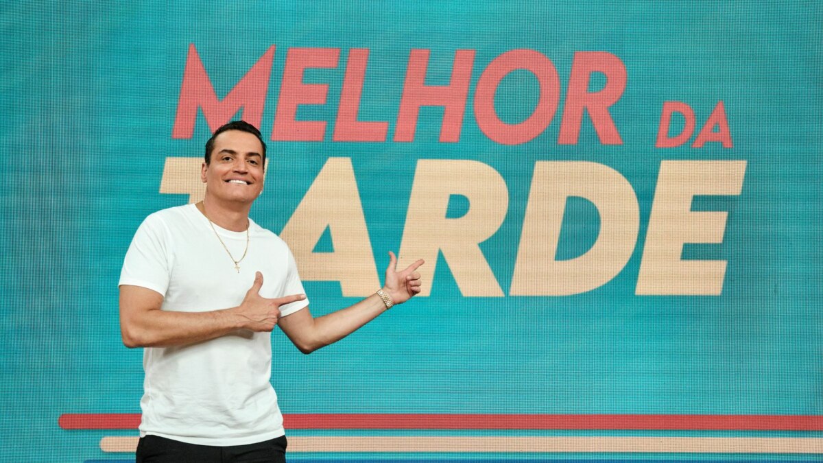 A imagem mostra o apresentador Léo Dias, sorrindo e usando uma camiseta branca e calças pretas. Ele aponta com as duas mãos para um painel com a inscrição "MELHOR DA TARDE" em letras maiúsculas vermelhas, sobre um fundo azul-petróleo. A foto foi tirada em um estúdio de televisão e a iluminação é clara.