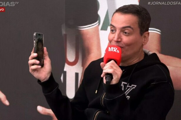 O apresentador Leo Dias, com um moletom preto, sorri enquanto segura um celular e um microfone. Ele está em um estúdio de televisão, e o nome do arquivo sugere que ele está ligando para Cariúcha.