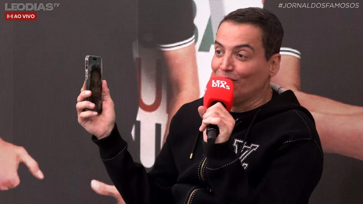 O apresentador Leo Dias, com um moletom preto, sorri enquanto segura um celular e um microfone. Ele está em um estúdio de televisão, e o nome do arquivo sugere que ele está ligando para Cariúcha.