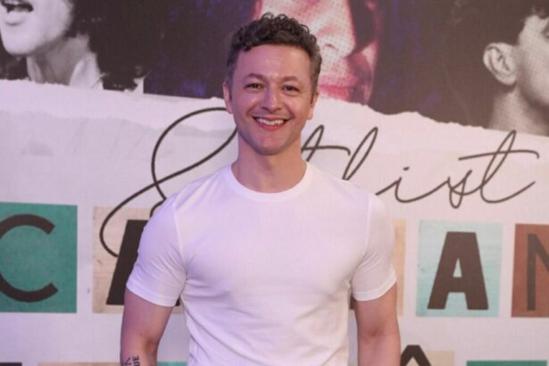 O músico Lucas Lima, em um close-up de meio-corpo, sorri para a câmera. Ele veste uma camiseta branca lisa. O fundo da foto é um banner com letras e imagens desfocadas.