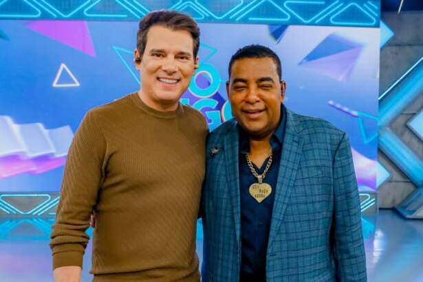 Celso Portiolli e Luiz Carlos, vocalista do Raça Negra, em um estúdio de TV. Celso Portiolli, à esquerda, usa uma blusa marrom de manga comprida e calças de sarja claras, enquanto Luiz Carlos, à direita, veste um terno xadrez azul com uma camisa escura e um colar dourado. Eles estão lado a lado, sorrindo e com os braços sobre os ombros um do outro. Ao fundo, o cenário do programa "Domingo Legal" é visível, com painéis digitais azuis e brancos e luzes de estúdio