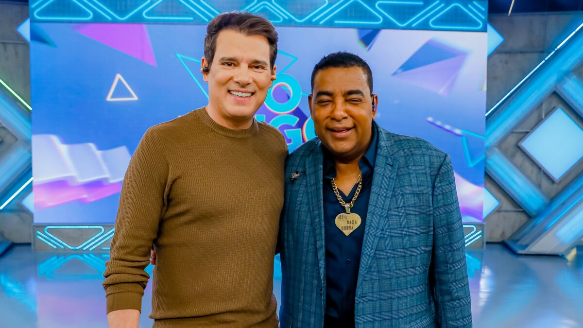 Celso Portiolli e Luiz Carlos, vocalista do Raça Negra, em um estúdio de TV. Celso Portiolli, à esquerda, usa uma blusa marrom de manga comprida e calças de sarja claras, enquanto Luiz Carlos, à direita, veste um terno xadrez azul com uma camisa escura e um colar dourado. Eles estão lado a lado, sorrindo e com os braços sobre os ombros um do outro. Ao fundo, o cenário do programa "Domingo Legal" é visível, com painéis digitais azuis e brancos e luzes de estúdio