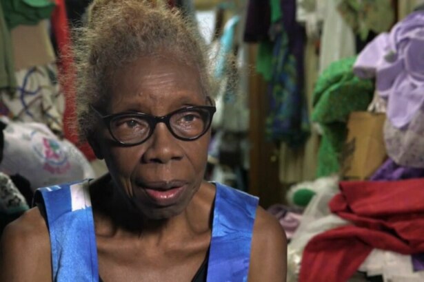 Mulher idosa de cabelos escuros e óculos, identificada como Maggie. Ela usa uma blusa azul estampada e olha para a câmera com uma expressão séria. O fundo está completamente bagunçado, cheio de roupas e objetos. O nome do arquivo a associa ao programa 'Acumuladores'.