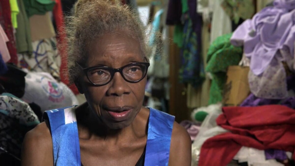 Mulher idosa de cabelos escuros e óculos, identificada como Maggie. Ela usa uma blusa azul estampada e olha para a câmera com uma expressão séria. O fundo está completamente bagunçado, cheio de roupas e objetos. O nome do arquivo a associa ao programa 'Acumuladores'.