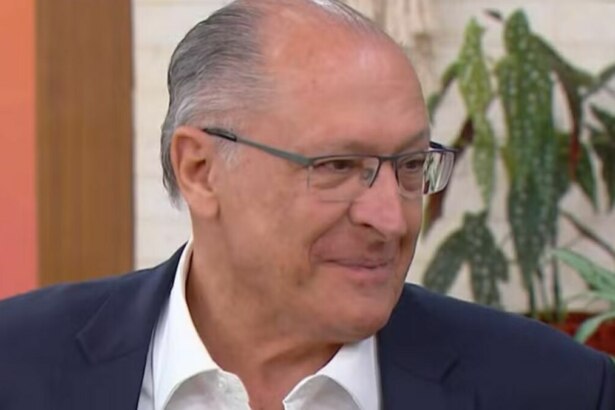 Um close-up de Geraldo Alckmin, um homem idoso de pele clara, com pouco cabelo grisalho penteado para trás. Ele usa óculos de armação fina e um terno azul marinho com uma camisa branca aberta no colarinho, em participação no Mais Você. Ele está olhando para a direita da imagem com uma expressão facial levemente sorridente e um fundo desfocado de plantas e objetos decorativos.