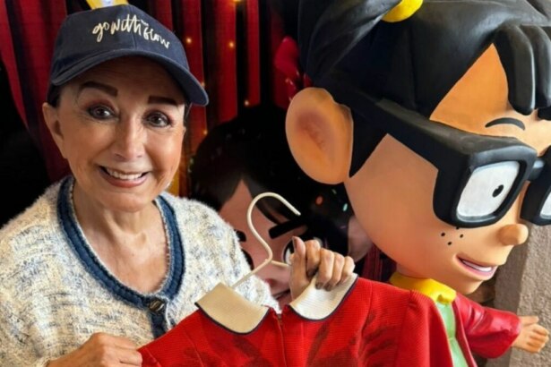 A imagem é um close da atriz Maria Antonieta de Las Nieves, conhecida por sua personagem Chiquinha do seriado "Chaves". Ela sorri para a câmera, usando um boné azul e um casaco de malha branco. Ela segura um cabide com uma roupa de cor vermelha. Ao lado dela, há uma grande estátua da personagem Chiquinha. O fundo é o cenário de um evento.