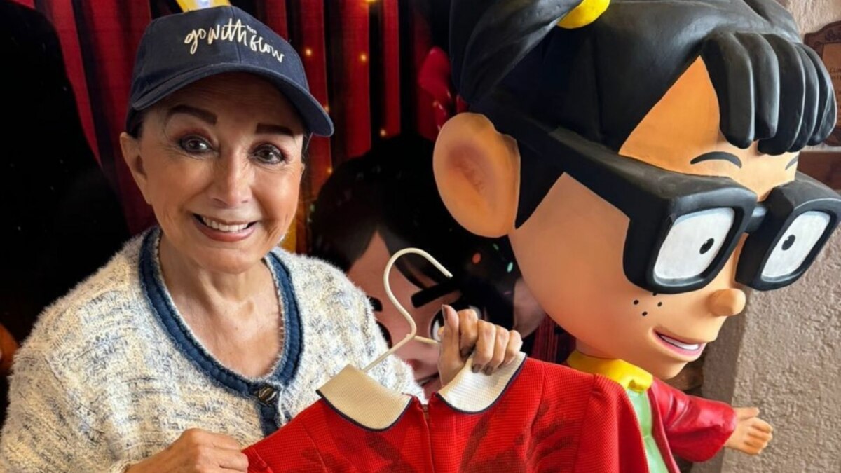 A imagem é um close da atriz Maria Antonieta de Las Nieves, conhecida por sua personagem Chiquinha do seriado "Chaves". Ela sorri para a câmera, usando um boné azul e um casaco de malha branco. Ela segura um cabide com uma roupa de cor vermelha. Ao lado dela, há uma grande estátua da personagem Chiquinha. O fundo é o cenário de um evento.