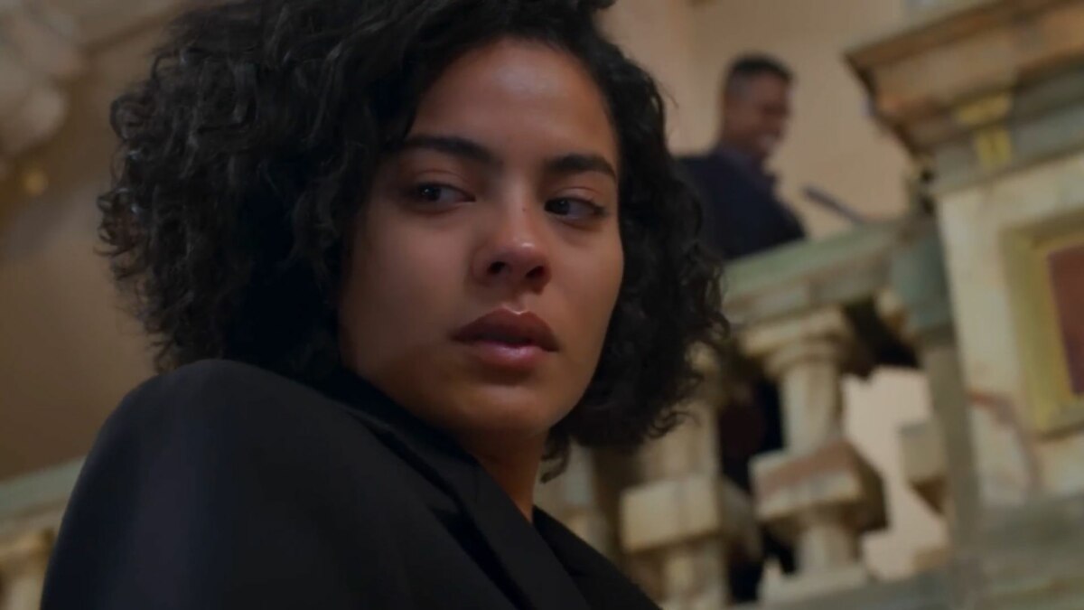 A imagem é um close da atriz Bella Campos, interpretando a personagem Maria de Fátima na novela "Vale Tudo". Ela tem cabelos curtos e cacheados, e sua expressão facial é séria, com o olhar desviado para o lado. Ela está usando uma jaqueta preta. Ao fundo, desfocado, é possível ver um homem em um ambiente interno, com colunas de pedra.
