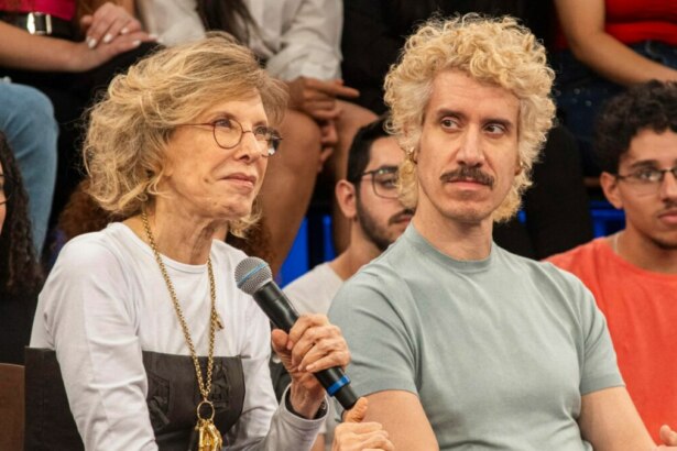 A apresentadora Marília Gabriela, com cabelo loiro curto e óculos de armação fina, segura um microfone e sorri discretamente. Ao lado dela, o filho Theodoro Cochrane, com cabelo loiro encaracolado e um bigode, olha seriamente para a frente. Ambos estão sentados em um estúdio com um público ao fundo.