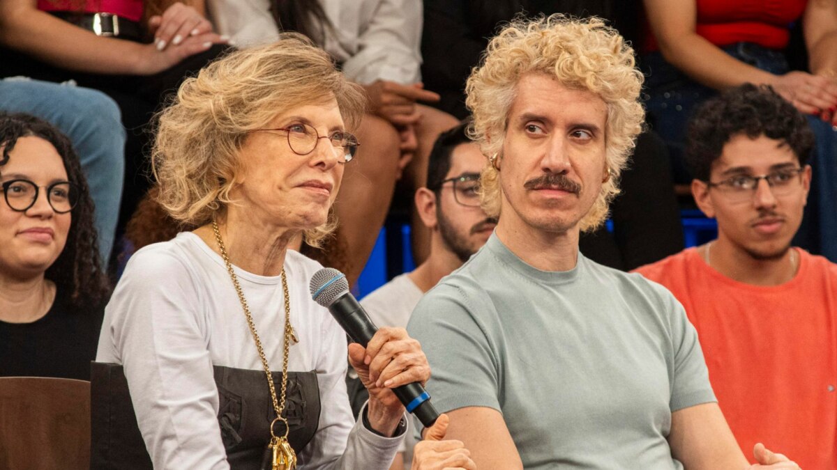 A apresentadora Marília Gabriela, com cabelo loiro curto e óculos de armação fina, segura um microfone e sorri discretamente. Ao lado dela, o filho Theodoro Cochrane, com cabelo loiro encaracolado e um bigode, olha seriamente para a frente. Ambos estão sentados em um estúdio com um público ao fundo.