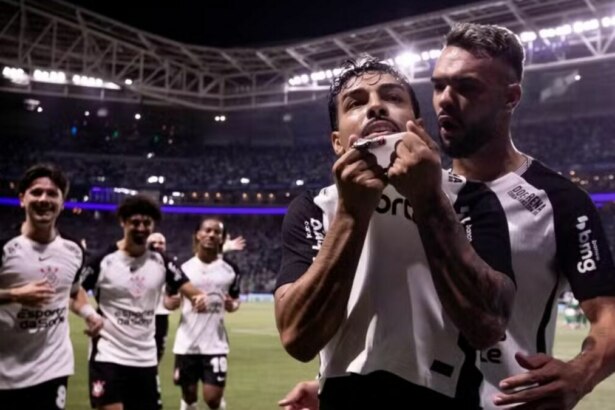 O jogador de futebol Matheus Bidu, do Corinthians, celebra um gol em um estádio lotado contra o Palmeiras. Ele puxa a camisa sobre o rosto e olha para cima, enquanto um colega de time o abraça. Outros jogadores do Corinthians correm para perto para comemorar, e as luzes do estádio iluminam a cena noturna.