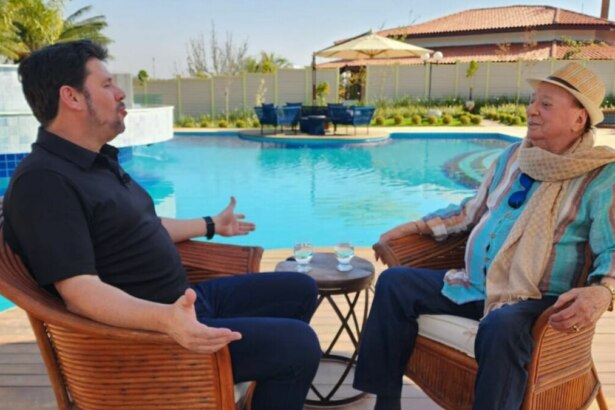 A imagem mostra uma cena do programa "Domingo Espetacular". O repórter Michael Keller e o apresentador Raul Gil estão sentados em cadeiras de vime, de frente um para o outro, em uma área externa com uma piscina. Michael Keller, à esquerda, veste uma camisa polo preta e calça escura, e gesticula com as mãos enquanto fala. Raul Gil, à direita, usa um chapéu de palha, uma camisa listrada e um cachecol. Entre eles, há uma pequena mesa com dois copos de água. O fundo é uma casa e uma área de lazer.