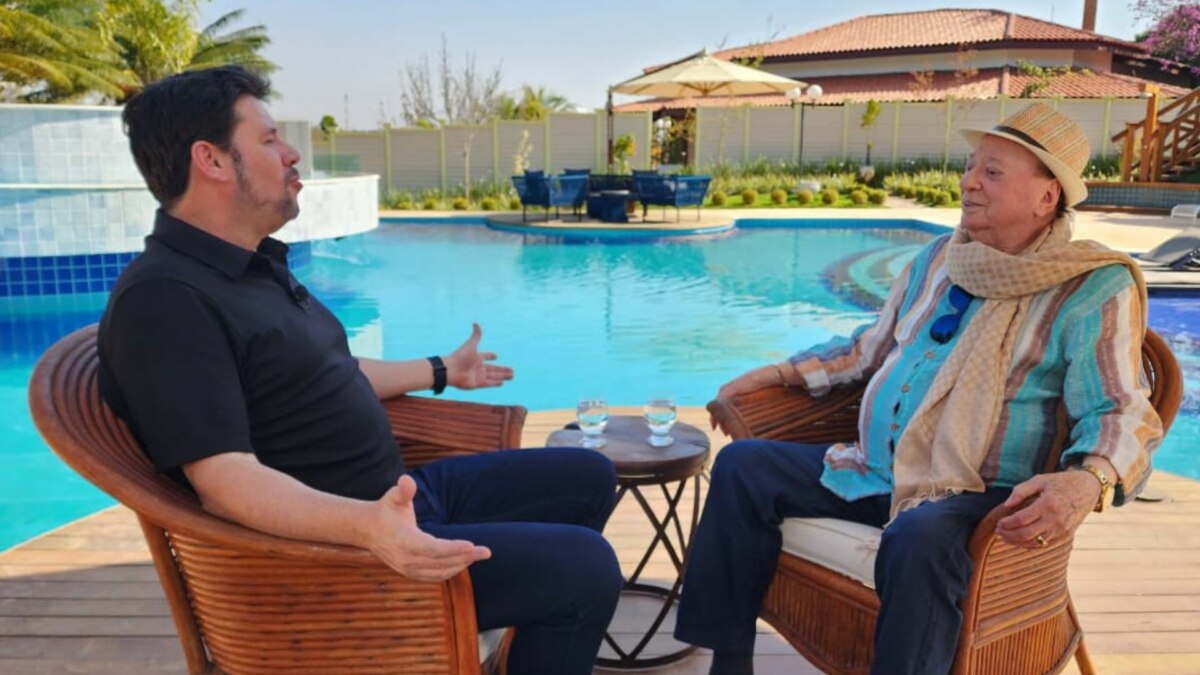 A imagem mostra uma cena do programa "Domingo Espetacular". O repórter Michael Keller e o apresentador Raul Gil estão sentados em cadeiras de vime, de frente um para o outro, em uma área externa com uma piscina. Michael Keller, à esquerda, veste uma camisa polo preta e calça escura, e gesticula com as mãos enquanto fala. Raul Gil, à direita, usa um chapéu de palha, uma camisa listrada e um cachecol. Entre eles, há uma pequena mesa com dois copos de água. O fundo é uma casa e uma área de lazer.