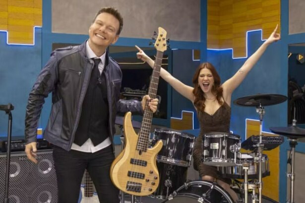 A imagem mostra Michel Teló e Ana Clara no cenário musical do programa Estrela da Casa. Michel Teló, à esquerda, sorri e segura um baixo elétrico. Ele usa uma jaqueta de couro azul-marinho, um colete preto e uma gravata. Ana Clara, à direita, está sentada atrás de uma bateria, com os braços erguidos em comemoração, e usa um vestido marrom. Ao fundo, o cenário é composto por formas geométricas em tons de azul e amarelo, com luzes de neon.