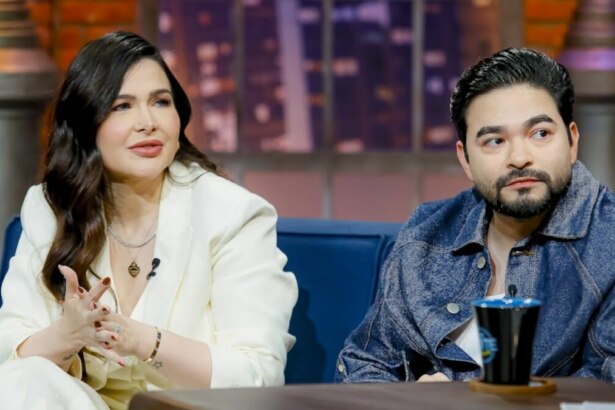 Mila Braga e Yudi Tamashiro sentados em um sofá azul no estúdio do programa 'The Noite'. Mila, de cabelo escuro, usa um blazer branco e está com as mãos unidas. Yudi, com barba e jaqueta jeans, também está sentado ao lado dela. Ambos parecem estar em uma entrevista.