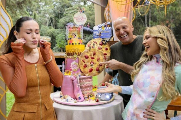 A apresentadora Nadja Haddad, à esquerda, com os jurados Giuseppe Gerundino e Beca Milano, em pé ao redor de um grande bolo temático com uma decoração que inclui uma pizza. Os três parecem rir da criação, em trecho da estreia do Bake Off Brasil 11