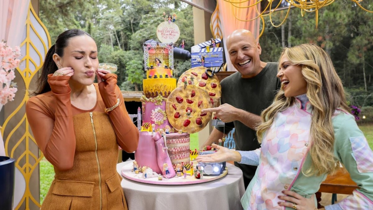 A apresentadora Nadja Haddad, à esquerda, com os jurados Giuseppe Gerundino e Beca Milano, em pé ao redor de um grande bolo temático com uma decoração que inclui uma pizza. Os três parecem rir da criação, em trecho da estreia do Bake Off Brasil 11