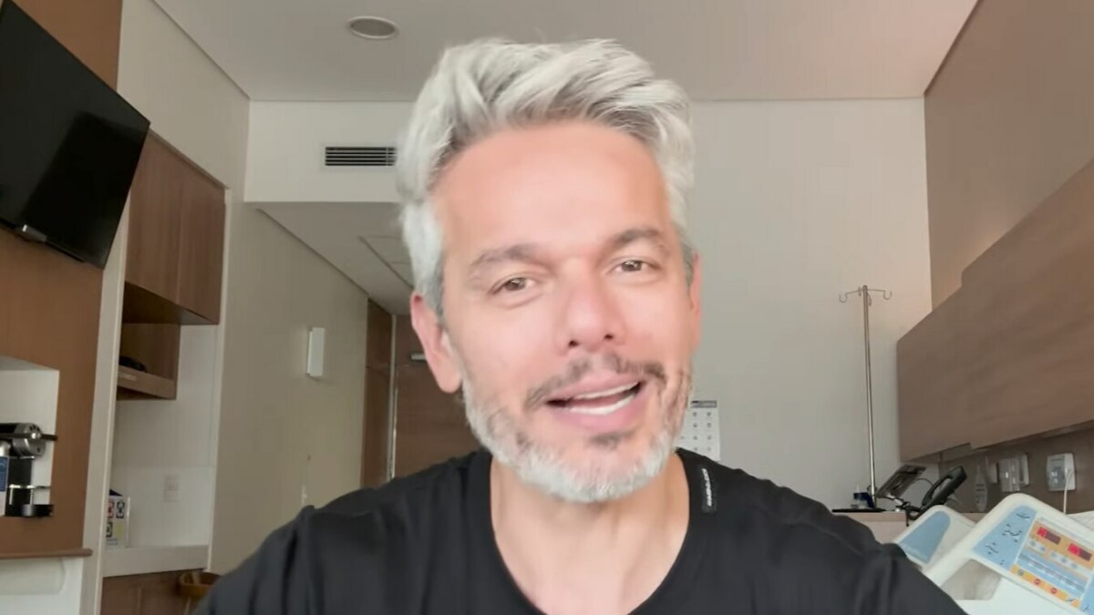 Otaviano Costa, com cabelo grisalho e barba, sorri em um quarto de hospital, após ser afastado do Melhor da Noite